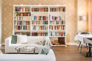 A living room with a white sofa, a glass coffee table, a bookshelf filled with books, and a dining area with a table and chairs. - AKZENT Hotel Dorn (Büsum)