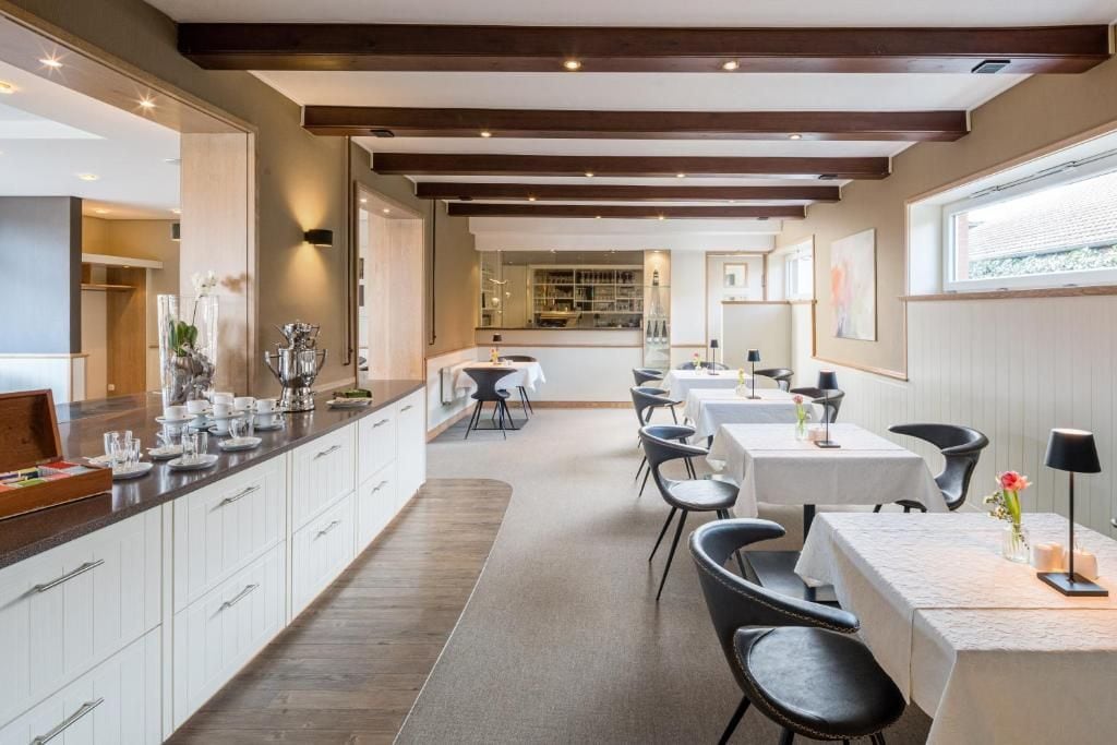 Et moderne spiseområde med en lang bar, hvide skabe og sorte stole. Der er dækket borde til spisning, og rummet har et træloft med synlige bjælker.
