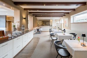 A modern dining area with a long counter, white cabinets, and black chairs. There are tables set for dining, and the space has a wooden ceiling with exposed beams. - AKZENT Hotel Dorn (Büsum)