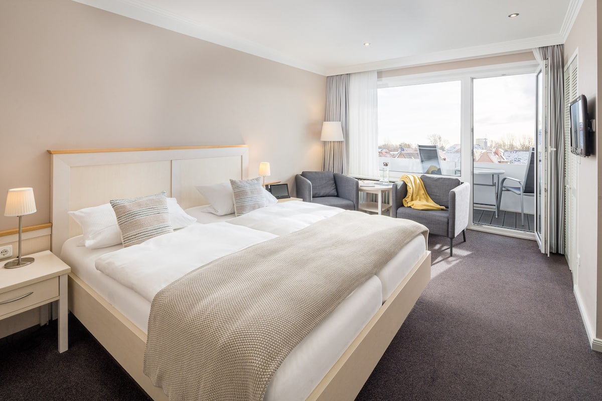 Habitación de hotel con una cama grande, un escritorio, una silla y vista a la ciudad por la ventana.