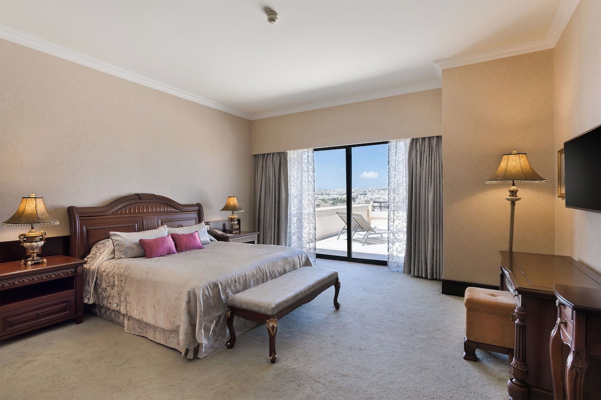 A hotel room with a large bed, a bench, two bedside tables with lamps, a TV, and a view of the city through large windows.