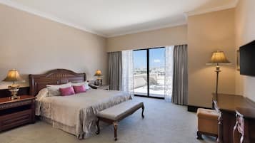 Habitación de hotel con una cama grande, un banco, dos mesitas de noche con lámparas, un televisor y vistas a la ciudad a través de grandes ventanales.