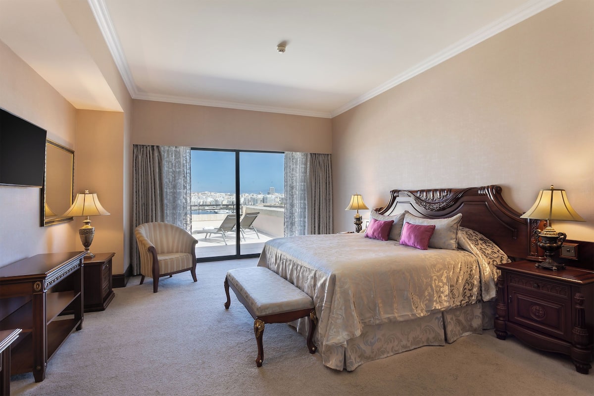 A spacious hotel room with a large bed, a chair, a desk, and a view of the city through large windows.