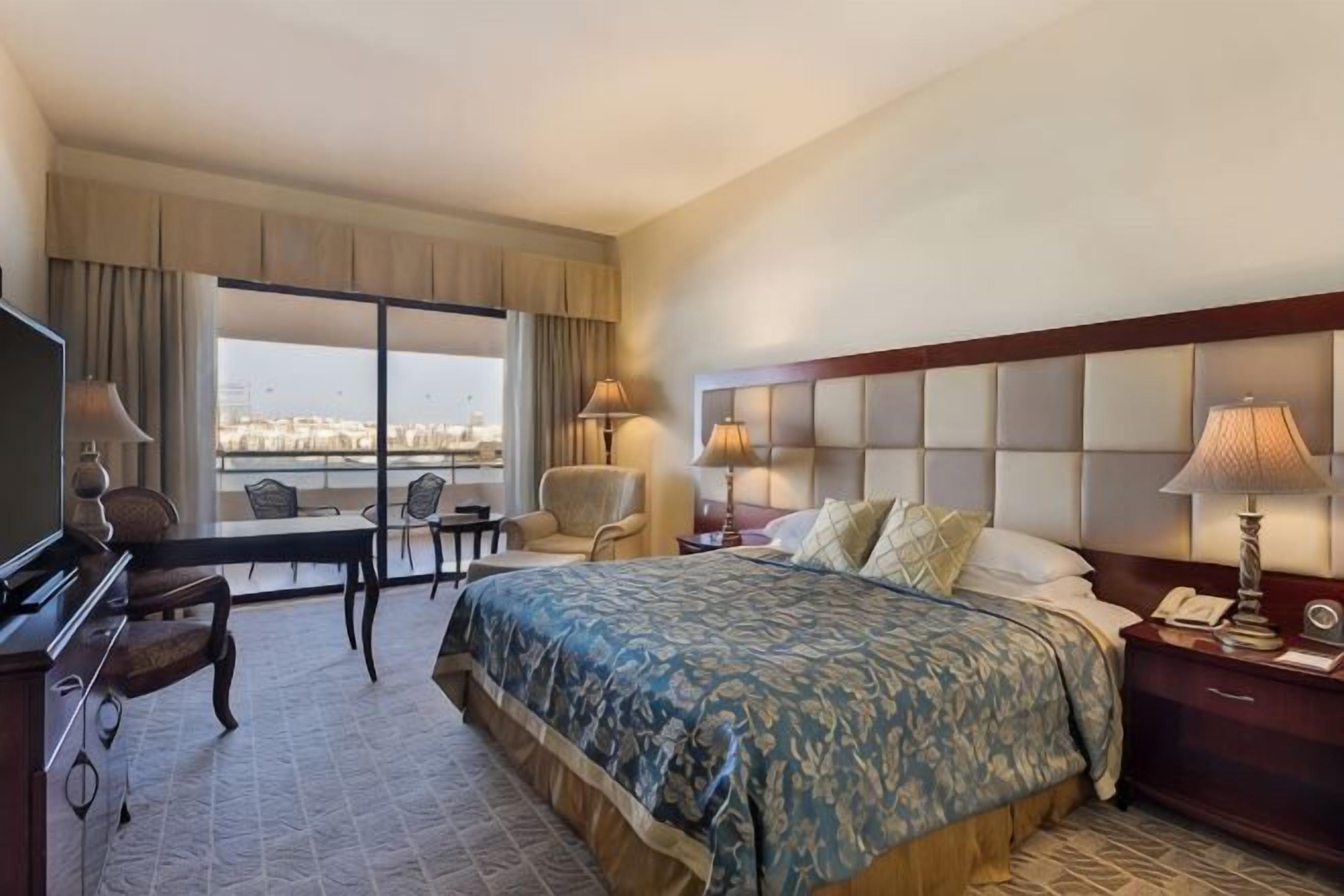 A hotel room with a large bed, a desk, a chair, a TV, a lamp, and a view of the city.