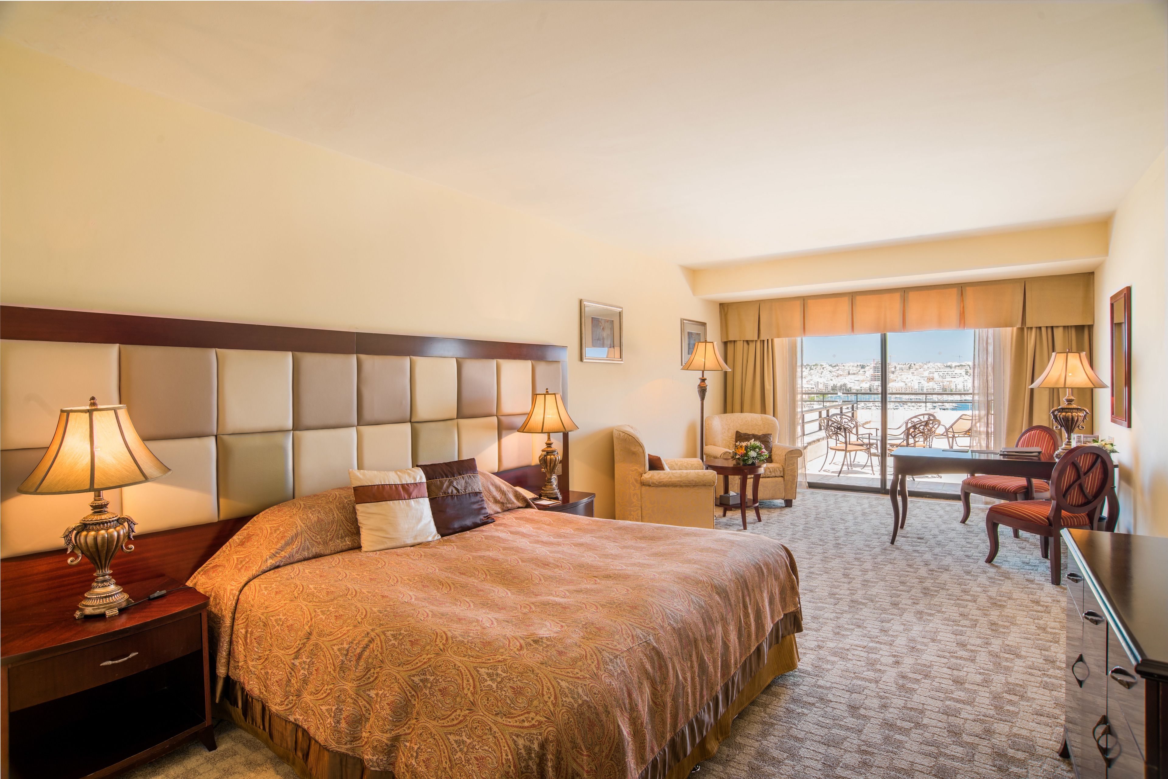 A hotel room with a large bed, two bedside tables with lamps, a small table with chairs, and a view of the city through large windows.