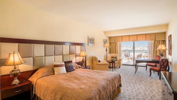Habitación de hotel con una cama grande, dos mesitas de noche con lámparas, una pequeña mesa con sillas y vistas a la ciudad a través de amplios ventanales.