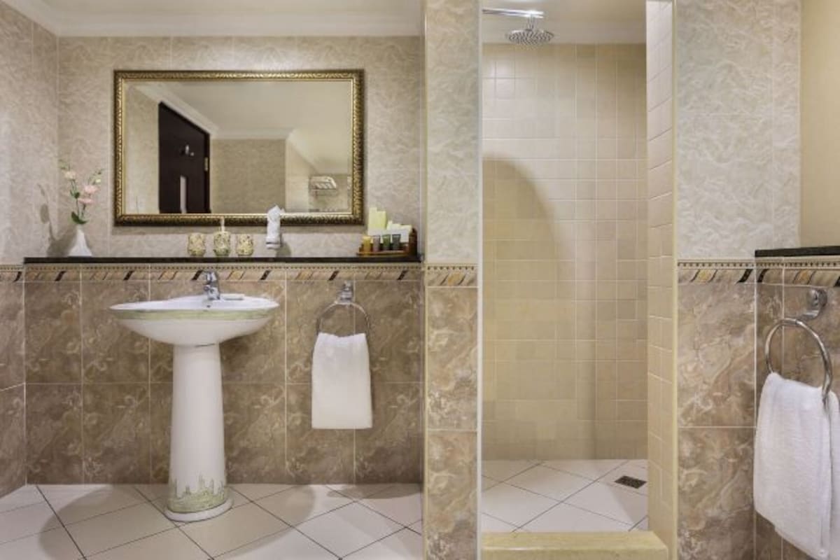 A bathroom with a pedestal sink, a mirror with a gold frame, a towel rack, and a shower area.