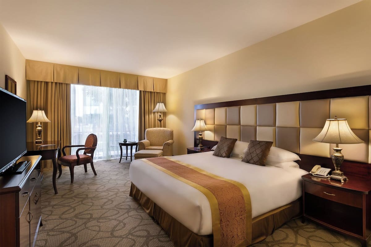 A hotel room with a large bed, two armchairs, a desk with a TV, and a window with curtains.