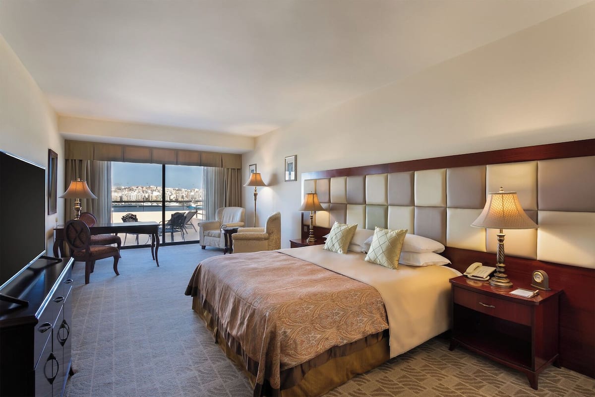 A hotel room with a large bed, a TV, a desk with a chair, and a view of the city through large windows.