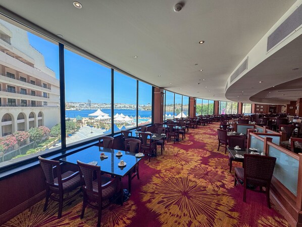 Un comedor amplio con ventanas grandes que ofrecen vistas a la ciudad y al mar. Las mesas están dispuestas en fila, y la habitación tiene un techo alto con iluminación empotrada.