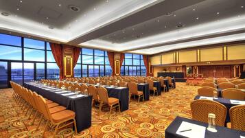 A conference room with rows of tables and chairs, large windows with curtains, and a patterned carpet.