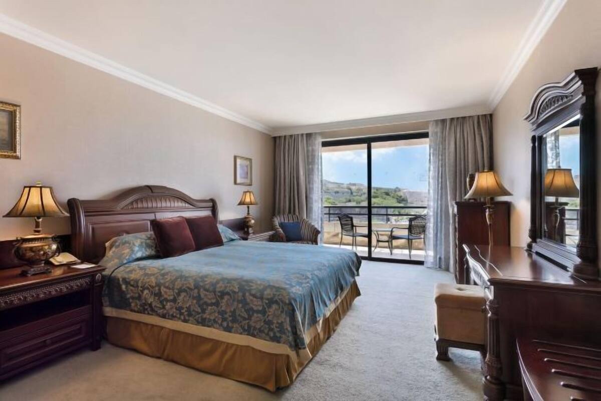 A hotel room with a large bed, a desk, a chair, a dresser, and a balcony with a view.