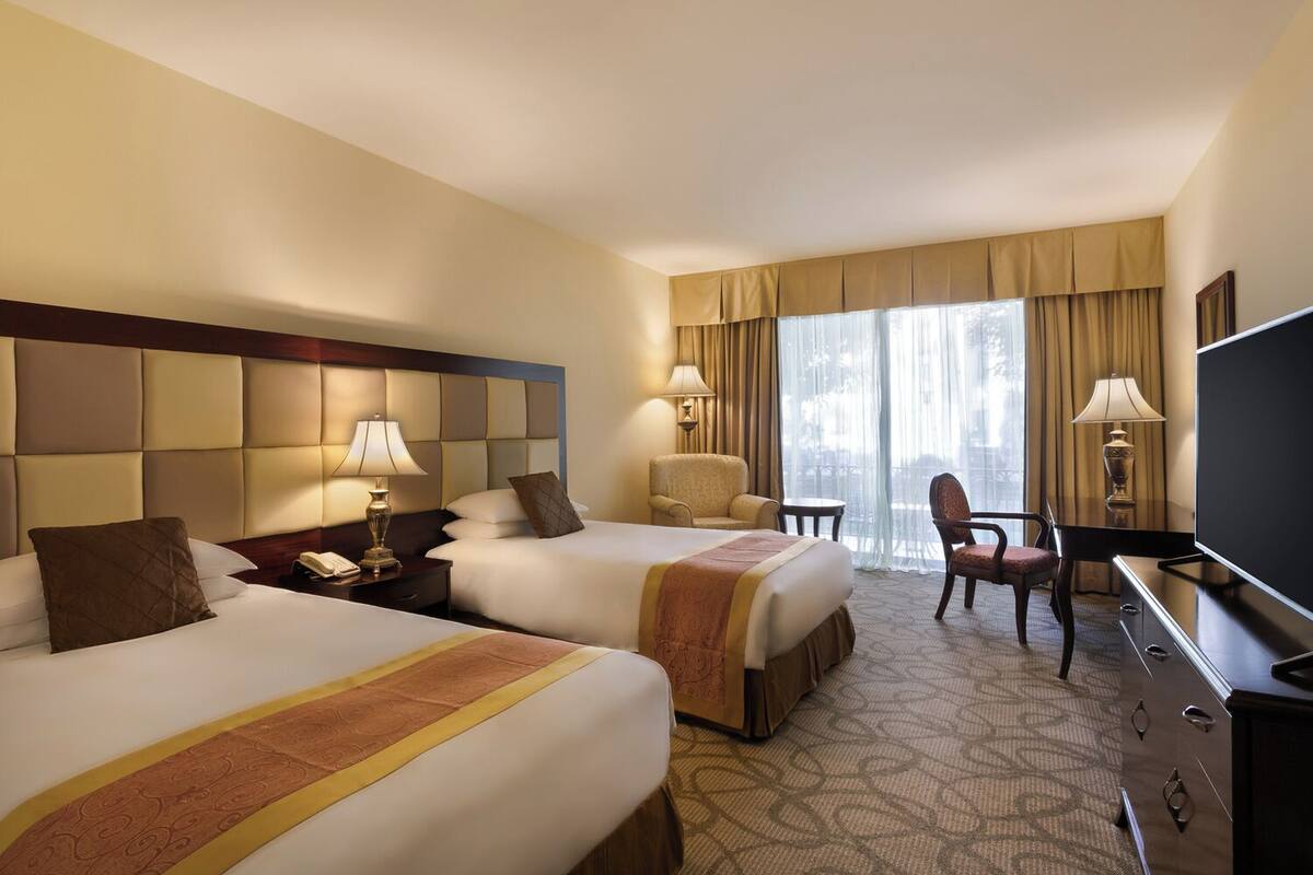 A hotel room with two beds, a desk, a chair, a television, and a window with curtains.