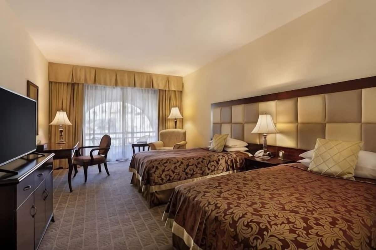 A hotel room with two beds, a desk with a TV, a chair, and a window with curtains.