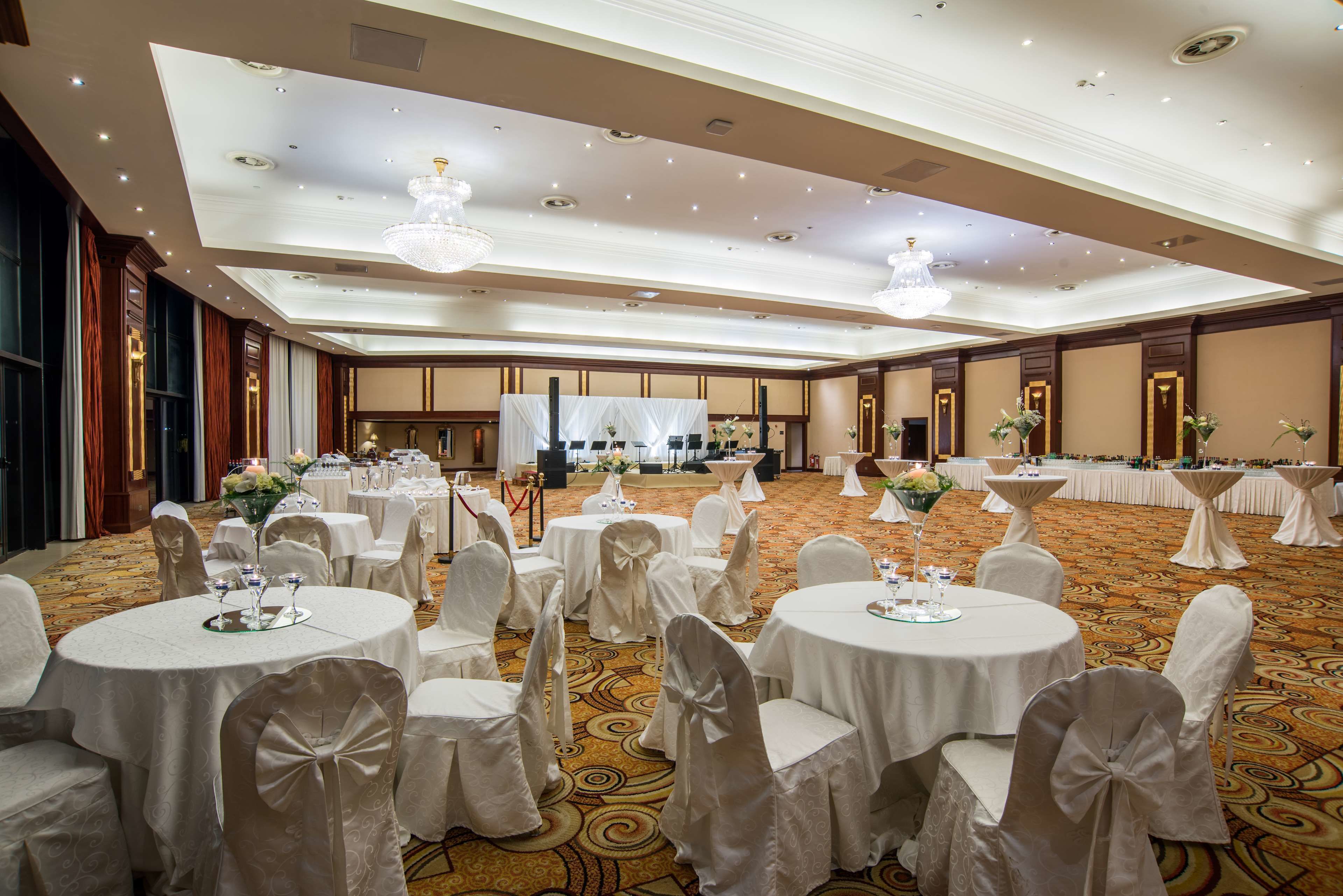 Een ruime balzaal met ronde tafels, witte stoelen en kroonluchters.