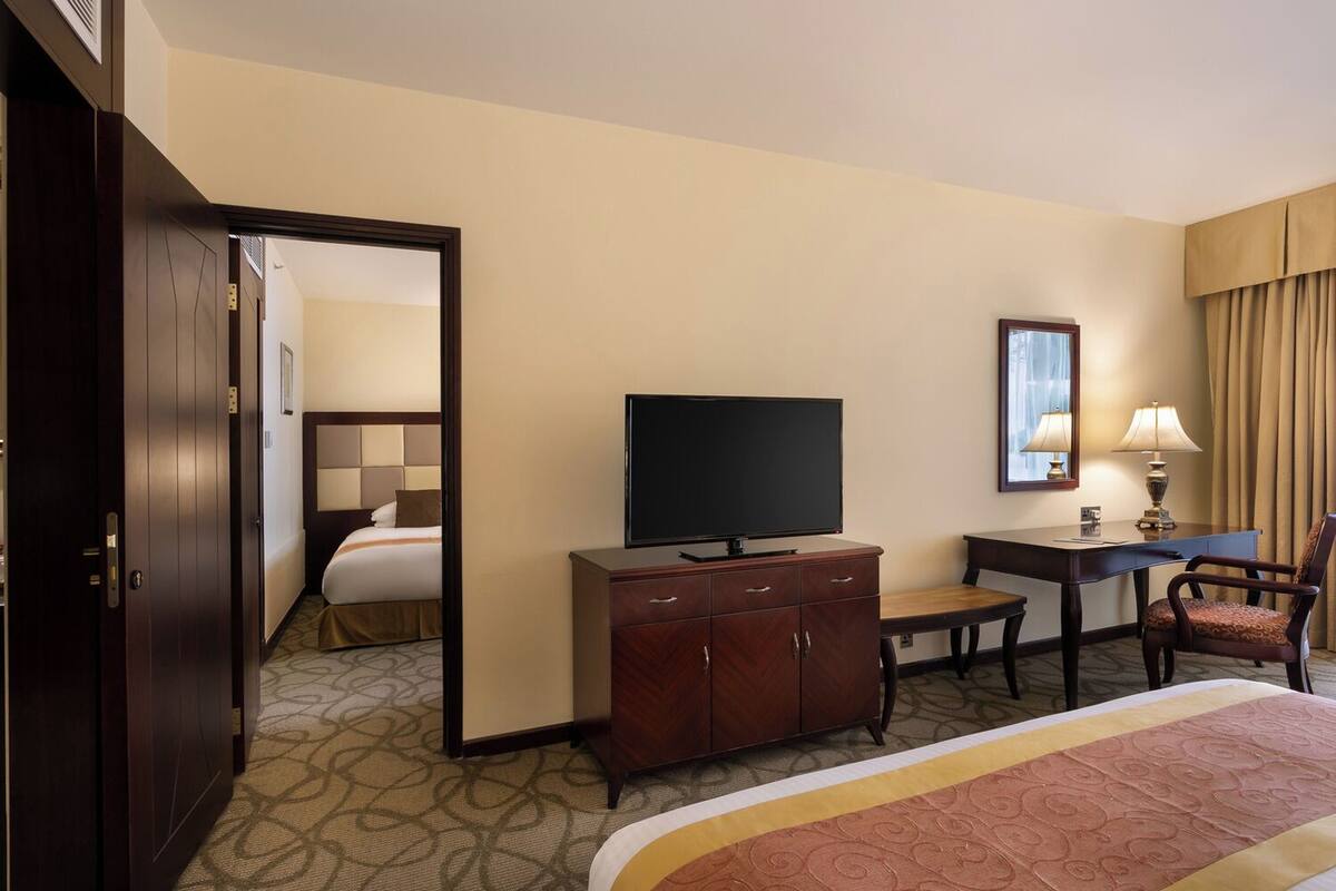 A hotel room with a bed, a TV on a wooden cabinet, a desk with a chair, and a mirror on the wall.