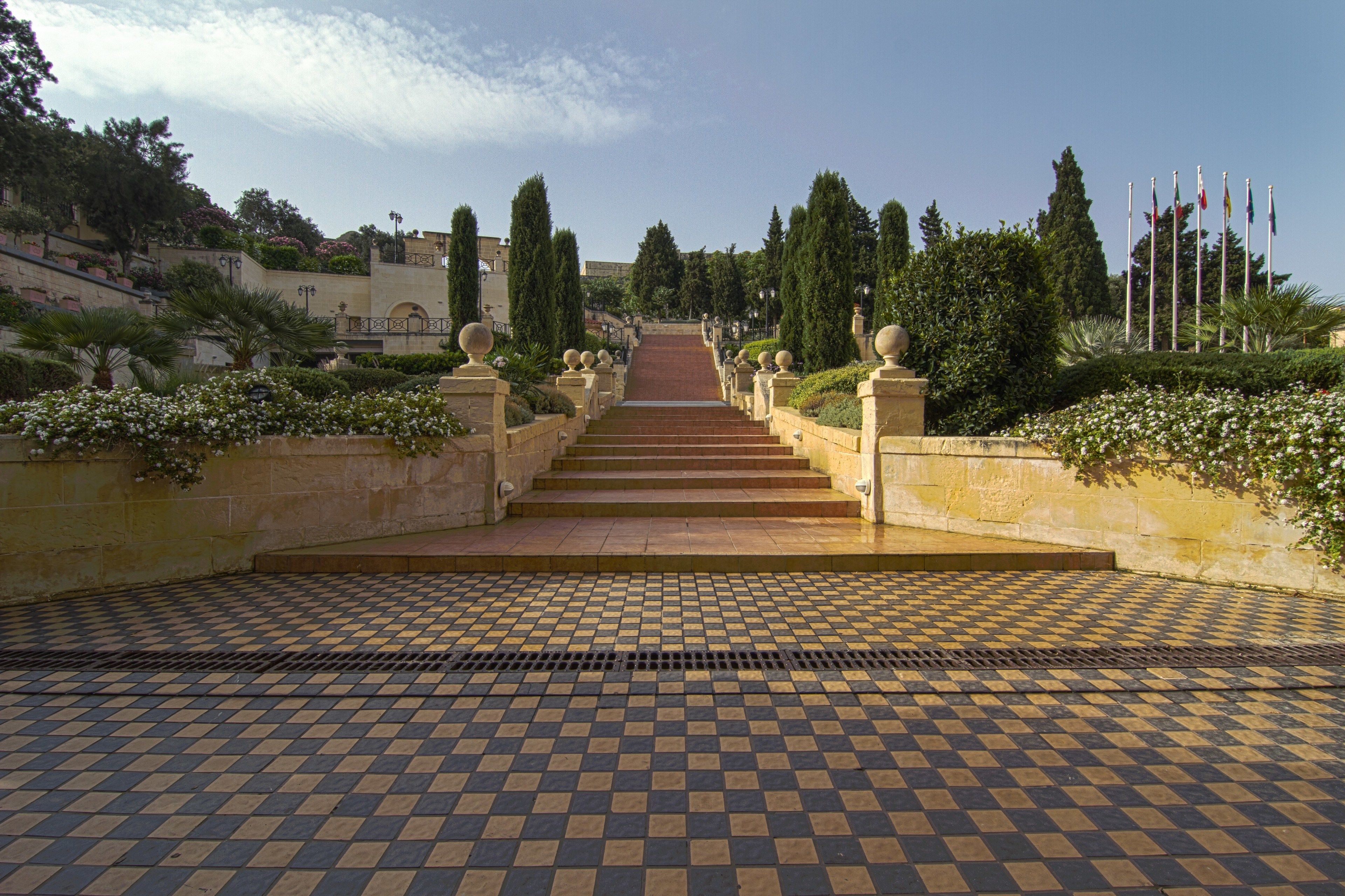 A checkered tile floor leading up a set of stairs with balustrades, surrounded by greenery and flags.