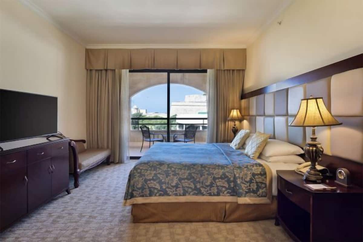 A hotel room with a large bed, a television, a desk with a lamp, and a view of the city through a large window.