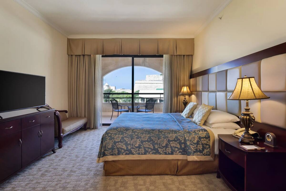 A hotel room with a large bed, a TV, a desk with a lamp, and a balcony with chairs.
