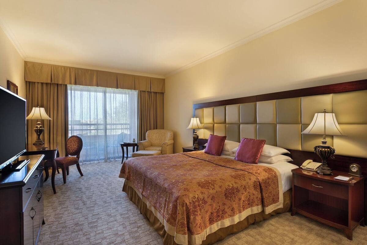 A hotel room with a large bed, a desk with a TV, a chair, and a nightstand with a lamp.