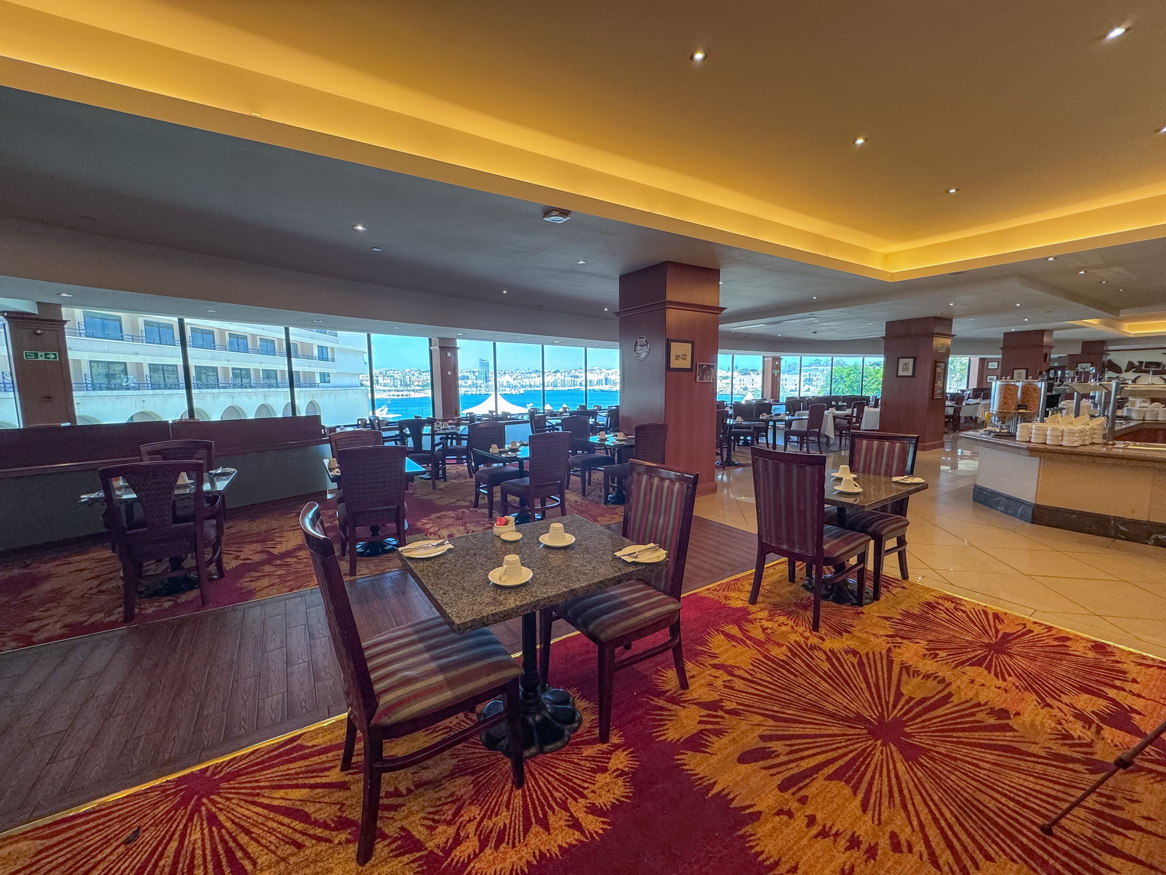 Un restaurante espacioso con techos altos, amplias ventanas que ofrecen vistas de la ciudad y diversas opciones de asientos, incluyendo mesas y sillas.