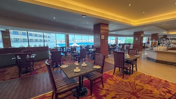 Un restaurante espacioso con techos altos, amplias ventanas que ofrecen vistas de la ciudad y diversas opciones de asientos, incluyendo mesas y sillas.