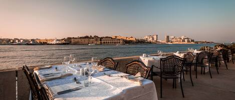 Un restaurante frente al mar con mesas dispuestas para comer, con vistas a un muelle histórico y un paisaje urbano.