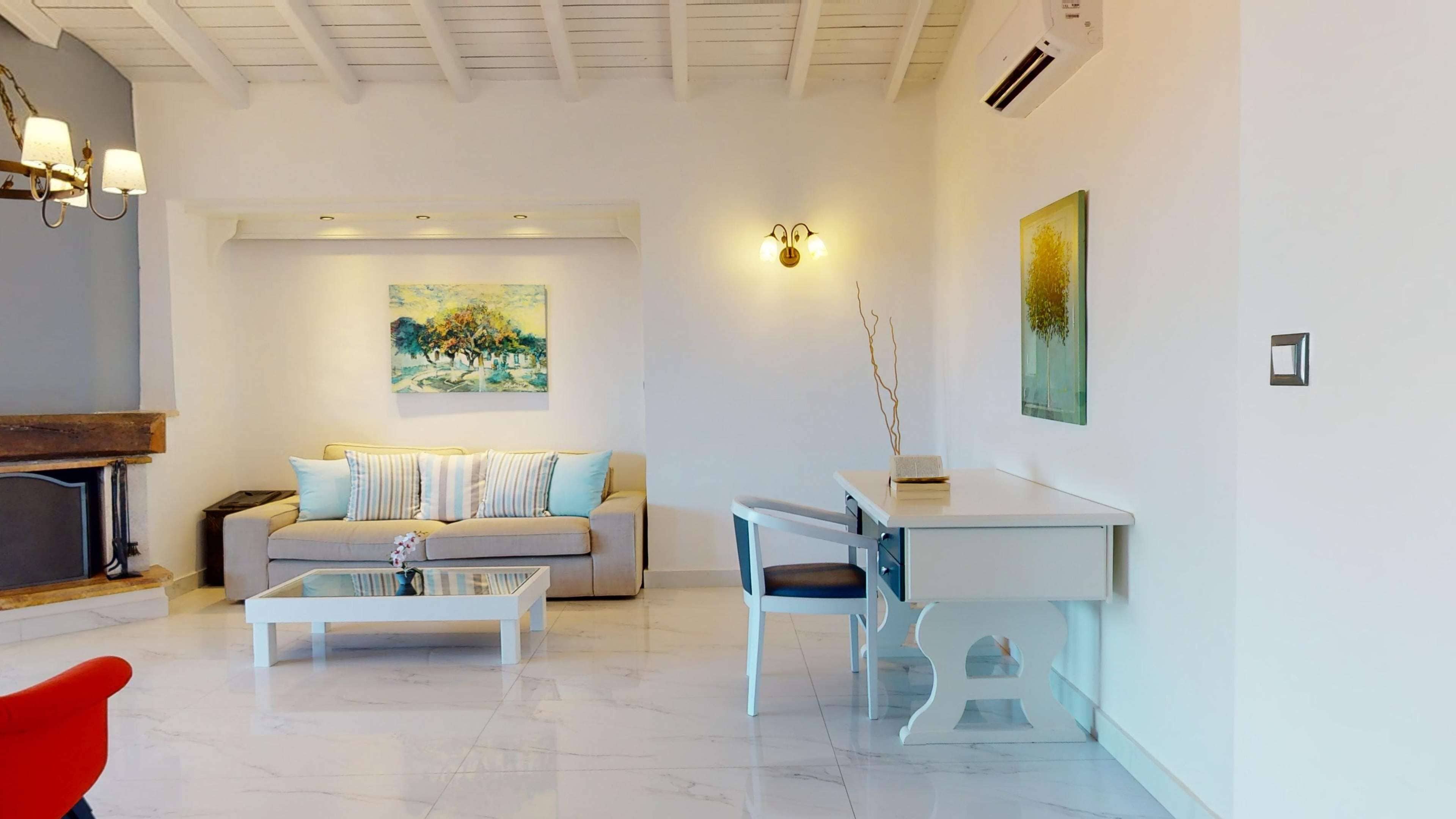 A modern living room with a sofa, coffee table, and a desk with a chair. There is a painting on the wall and an air conditioning unit.