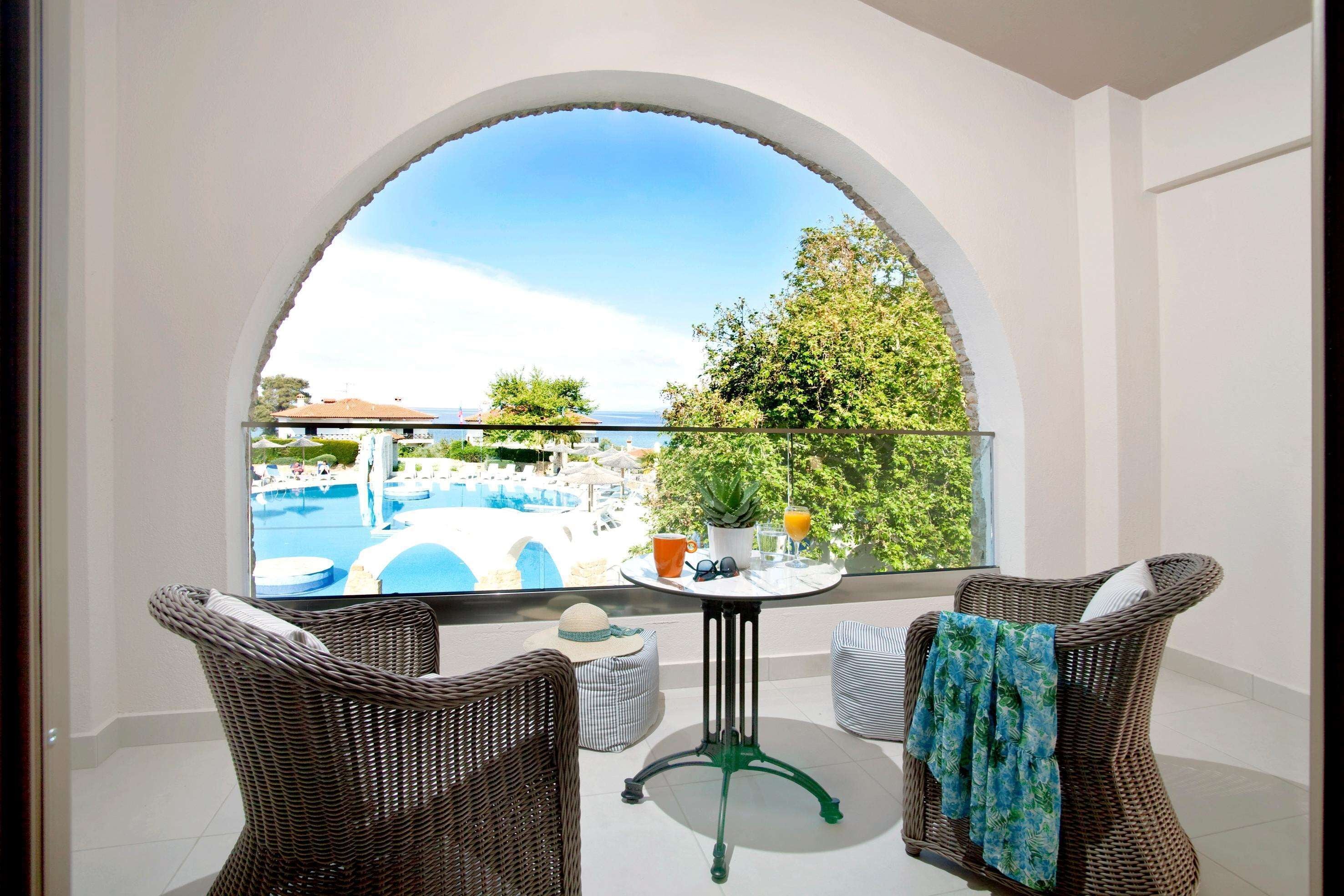 A balcony with wicker chairs, a small table with a potted plant, and a view of a swimming pool and greenery.