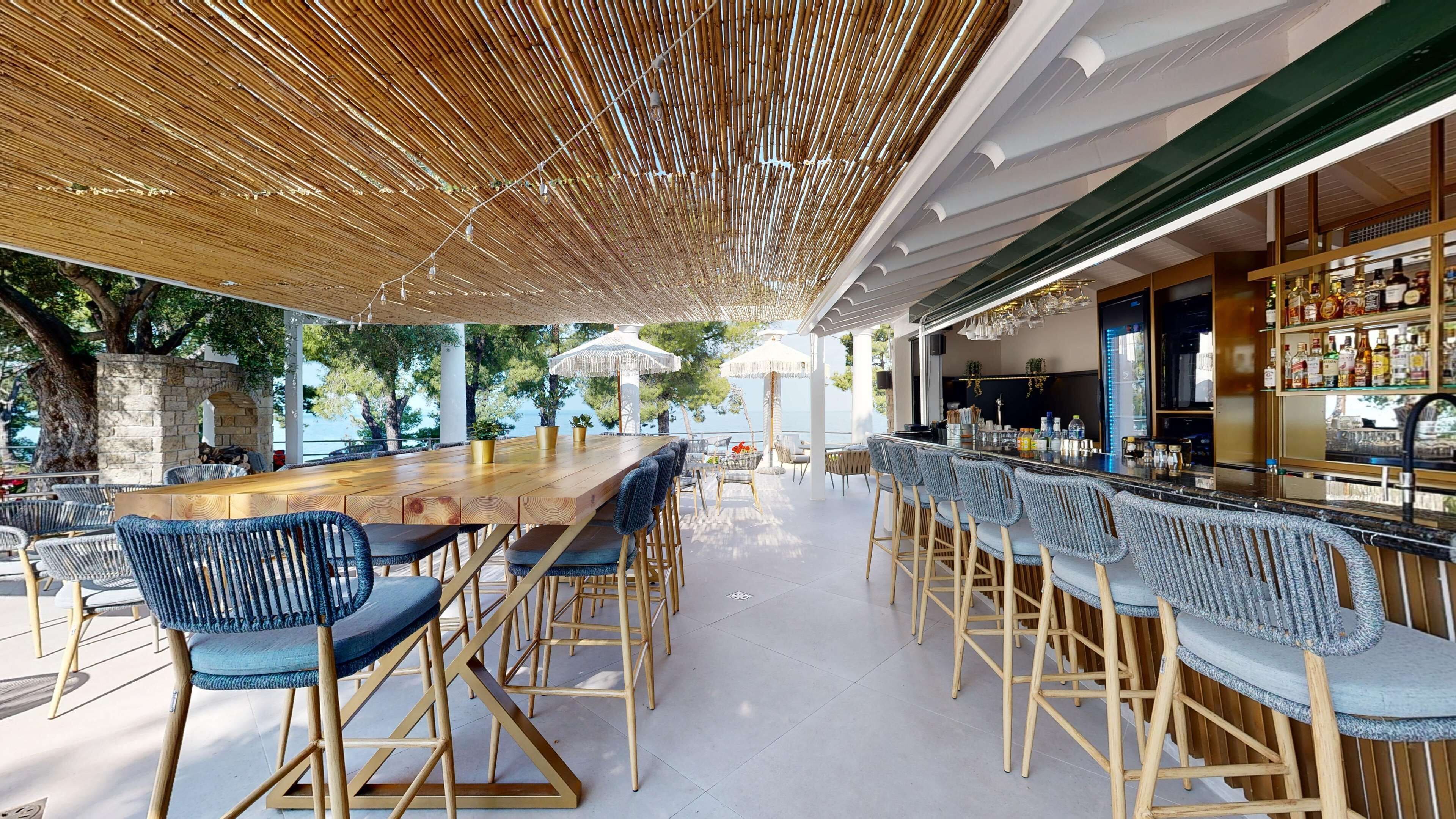 Un área de bar al aire libre con una larga mesa de madera y sillas tapizadas en azul, rodeada por un techo de bambú y con vistas panorámicas.