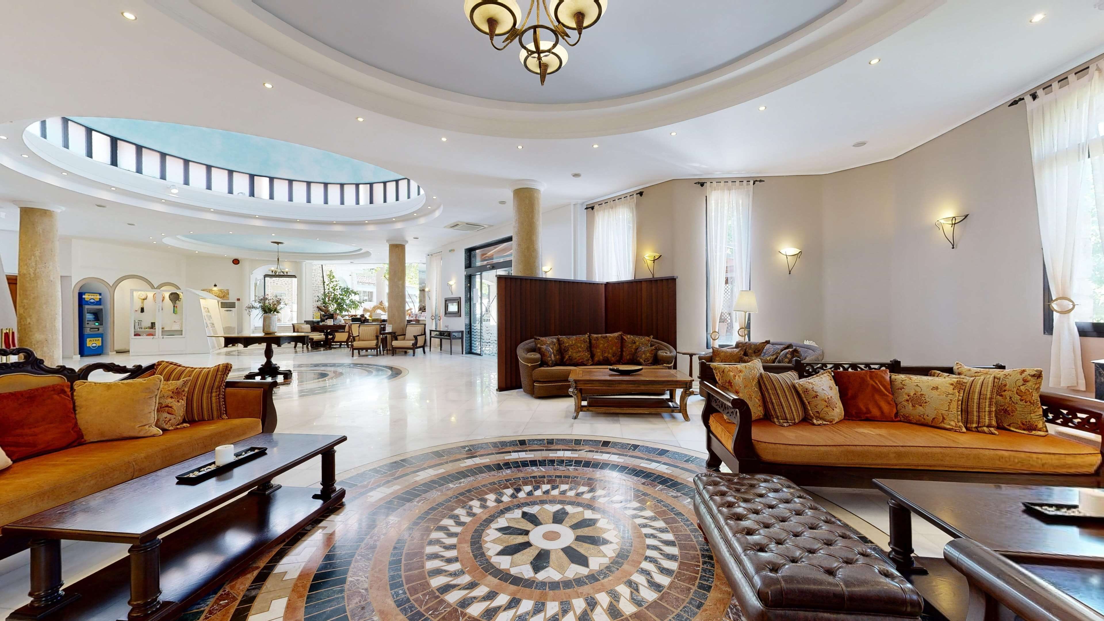 Um hall amplo com um chão em mosaico circular central, teto alto com lustre e área de estar com sofás e poltronas.