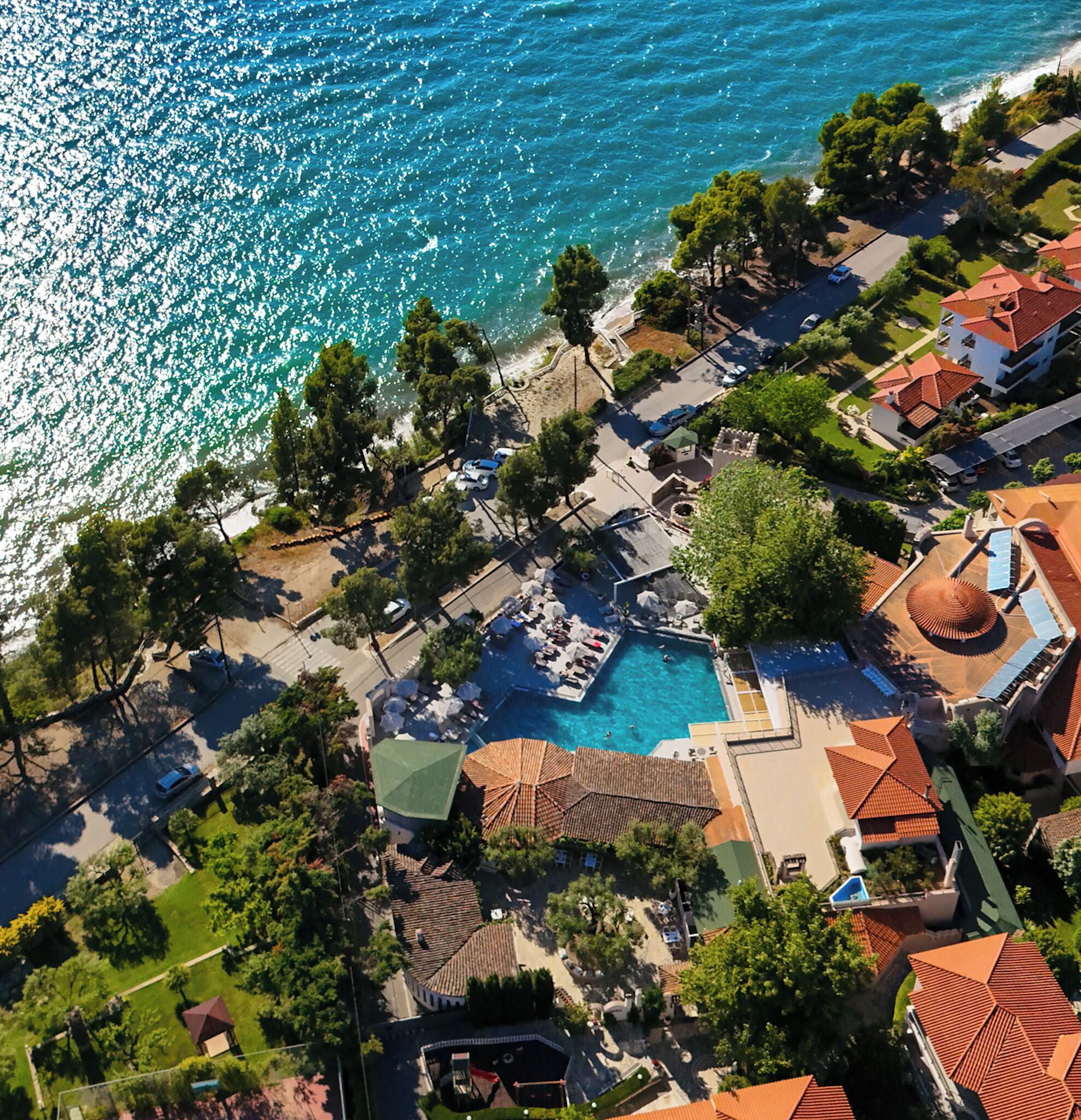 An aerial view of a coastal resort with a central swimming pool surrounded by lounge areas and a beach.