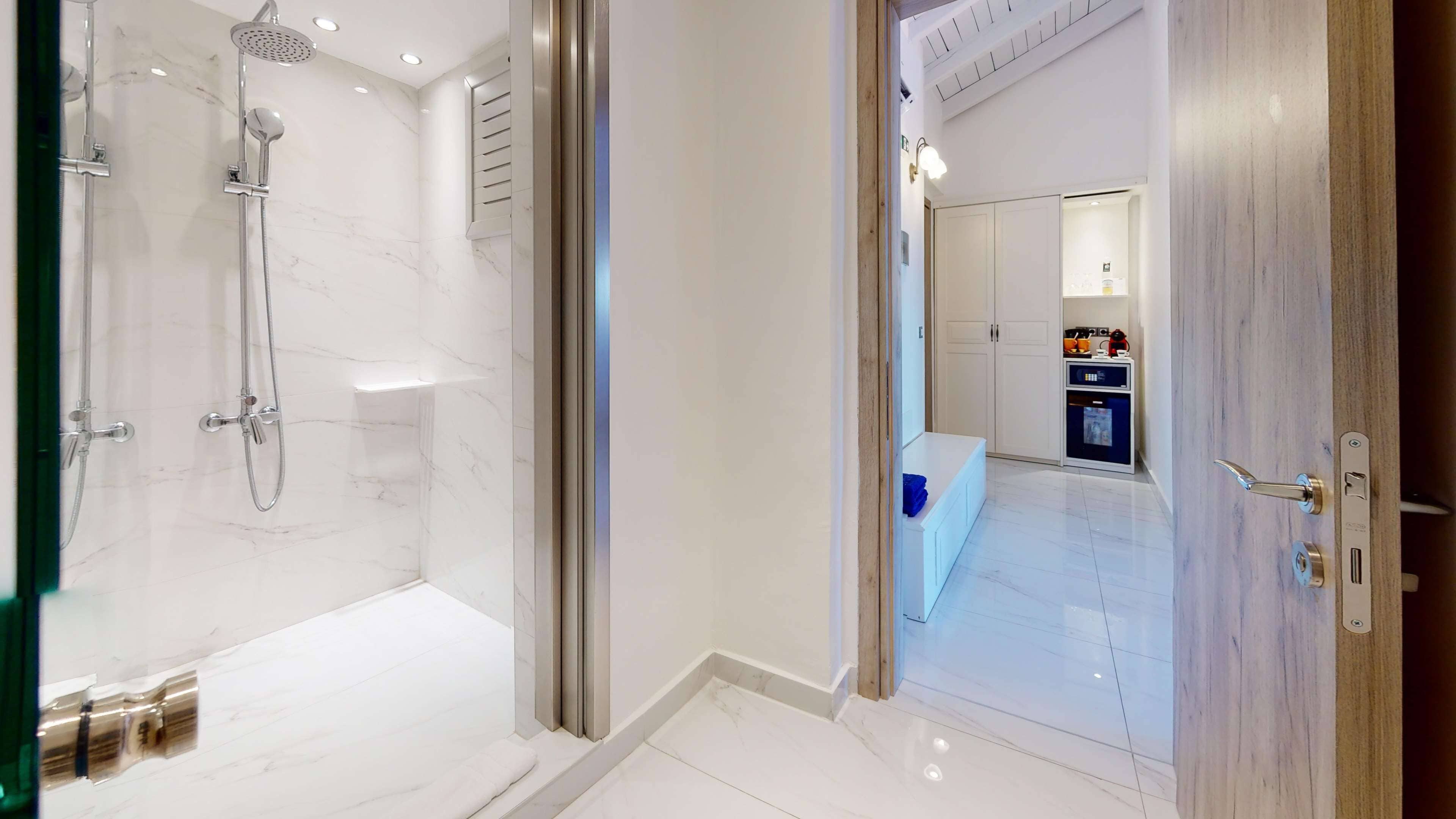 Un baño moderno con ducha envuelta en vidrio, suelo de mármol y puerta de madera que da a una zona de cocina.