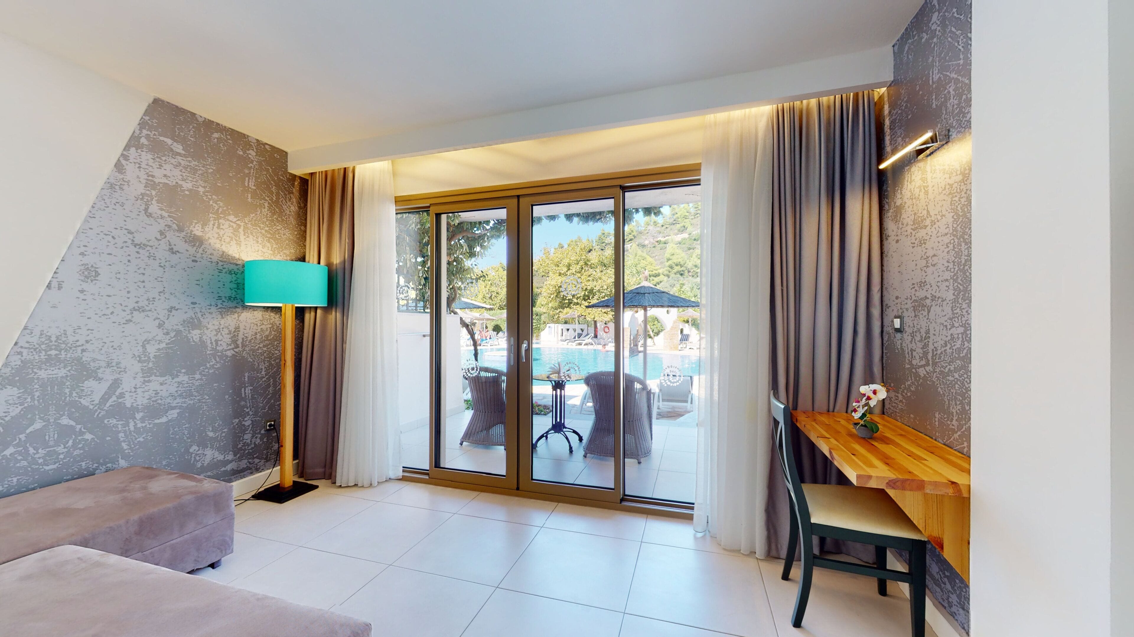 A modern hotel room with a large sliding glass door leading to a pool area, a wooden table with a vase of flowers, and a teal floor lamp.