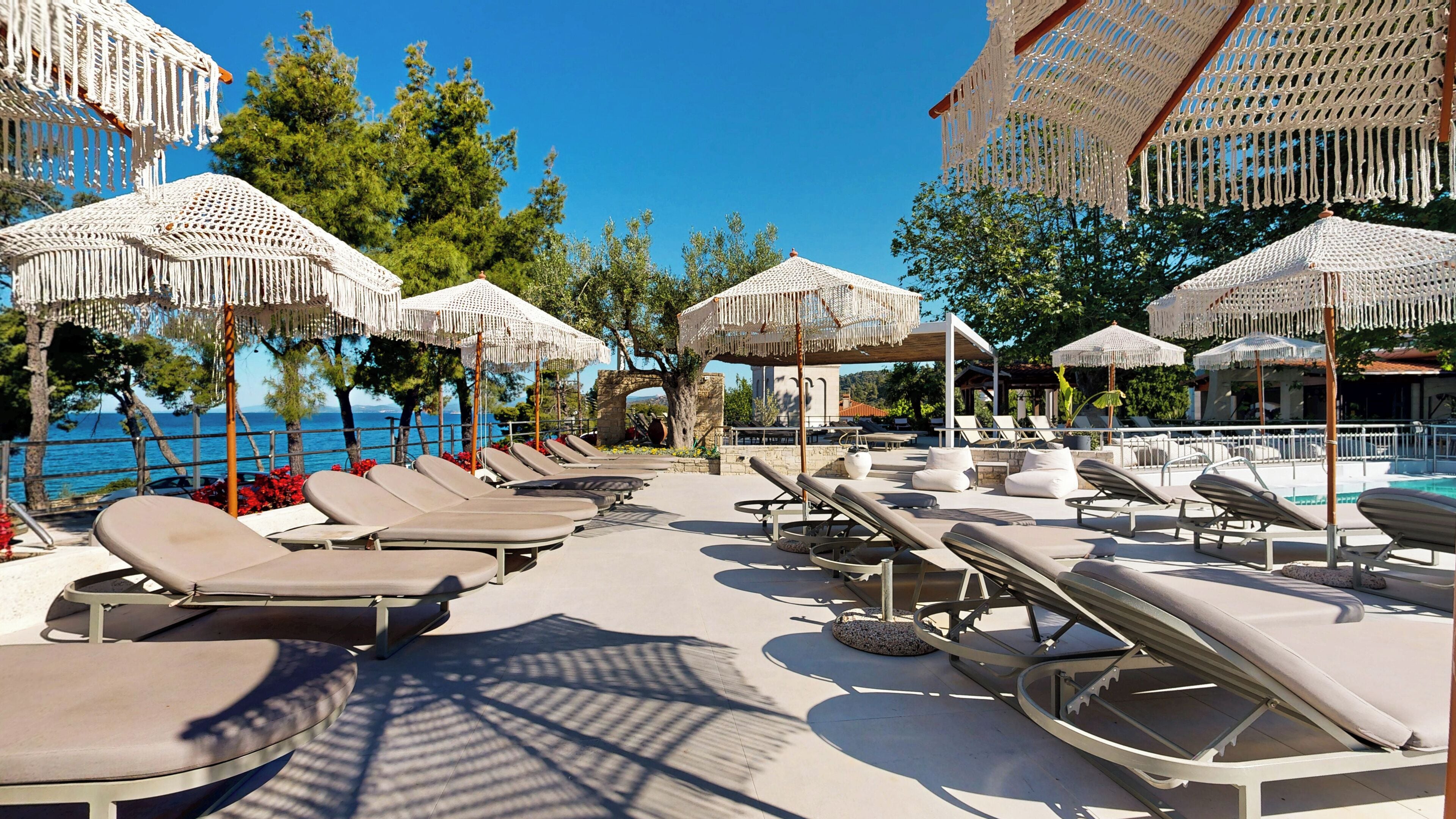 A poolside area with sun loungers and macrame umbrellas, overlooking a body of water.