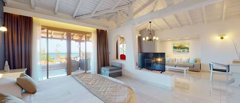 A spacious bedroom with a large bed, a balcony with curtains, a TV, and a seating area with a sofa and a coffee table.