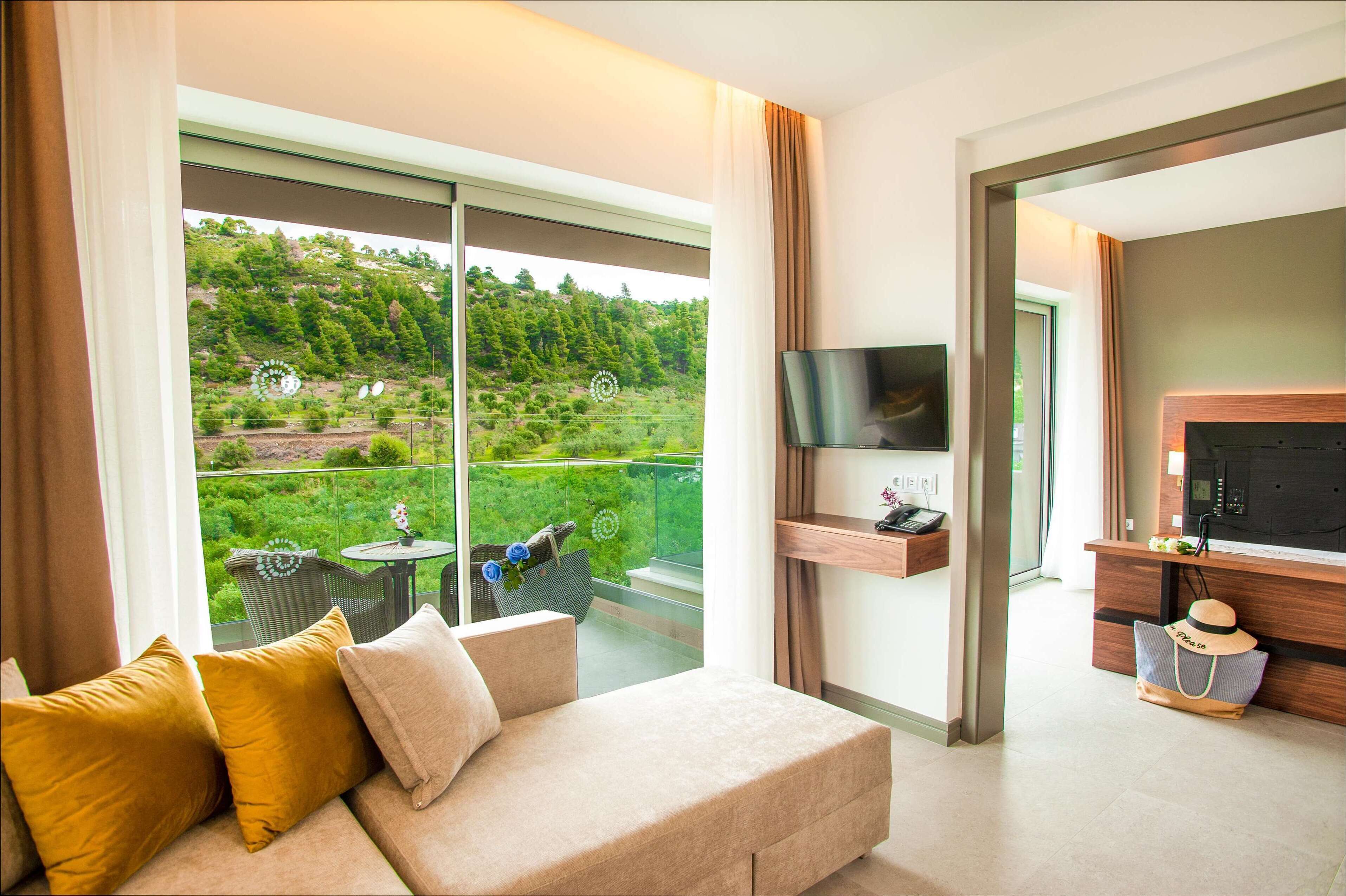 Ein modernes Hotelzimmer mit Balkon, einer Couch mit Kissen, einem Flachbildfernseher, einem Holztischchen und Blick auf eine grüne Landschaft.