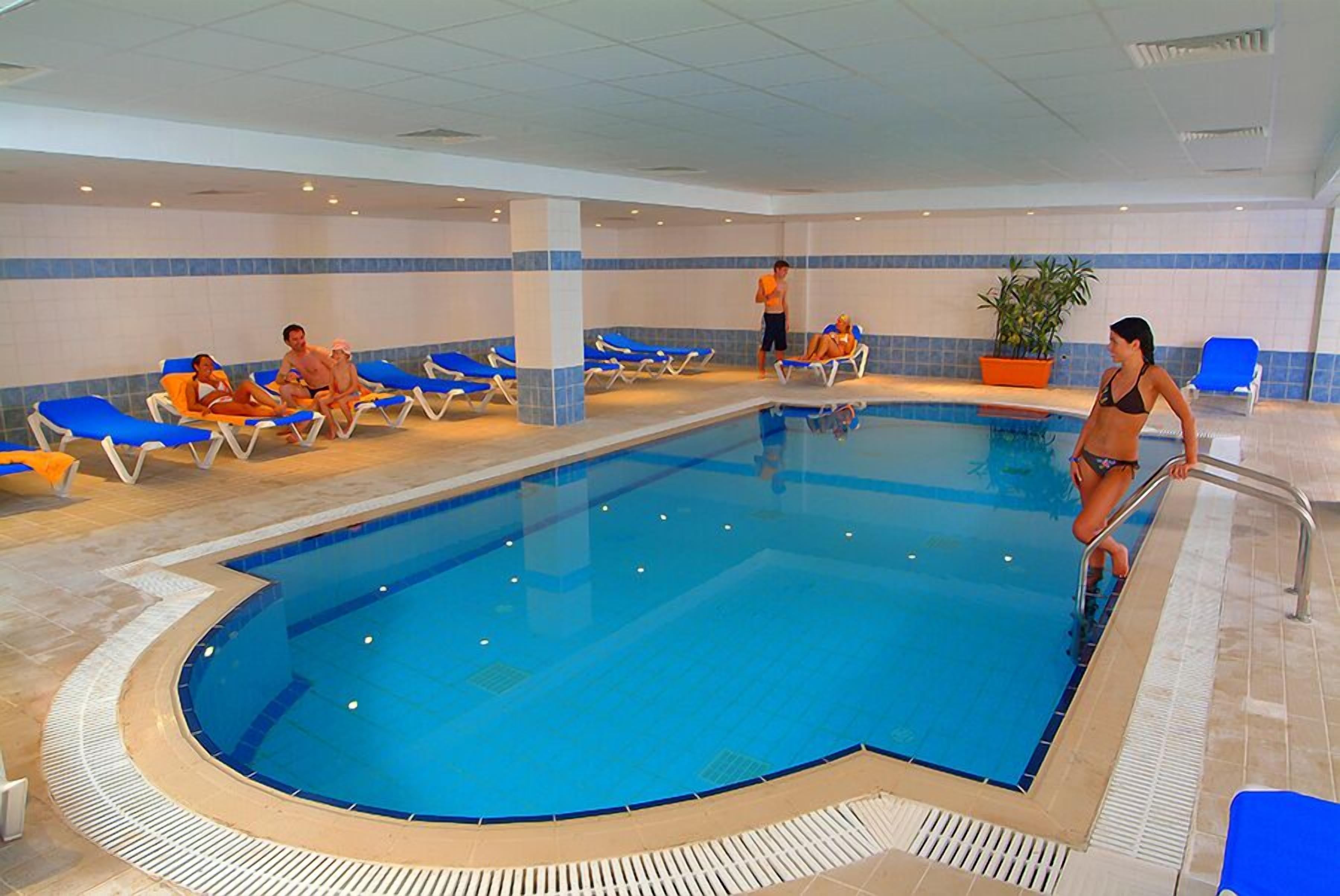 An indoor swimming pool with blue and white tiled walls, a pool with clear blue water, and several lounge chairs around the pool area.
