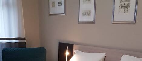 A double bed with white linens, a bedside table with a lamp, and framed pictures on the wall.