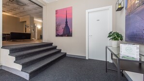 A hotel lobby with a staircase, a painting of a tower, a door, and a reception desk with a plant. - Hotel Gran Torino (Turin)