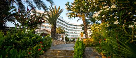 A hotel entrance with a stone staircase leading up to a curved building, surrounded by lush greenery and flowering plants.