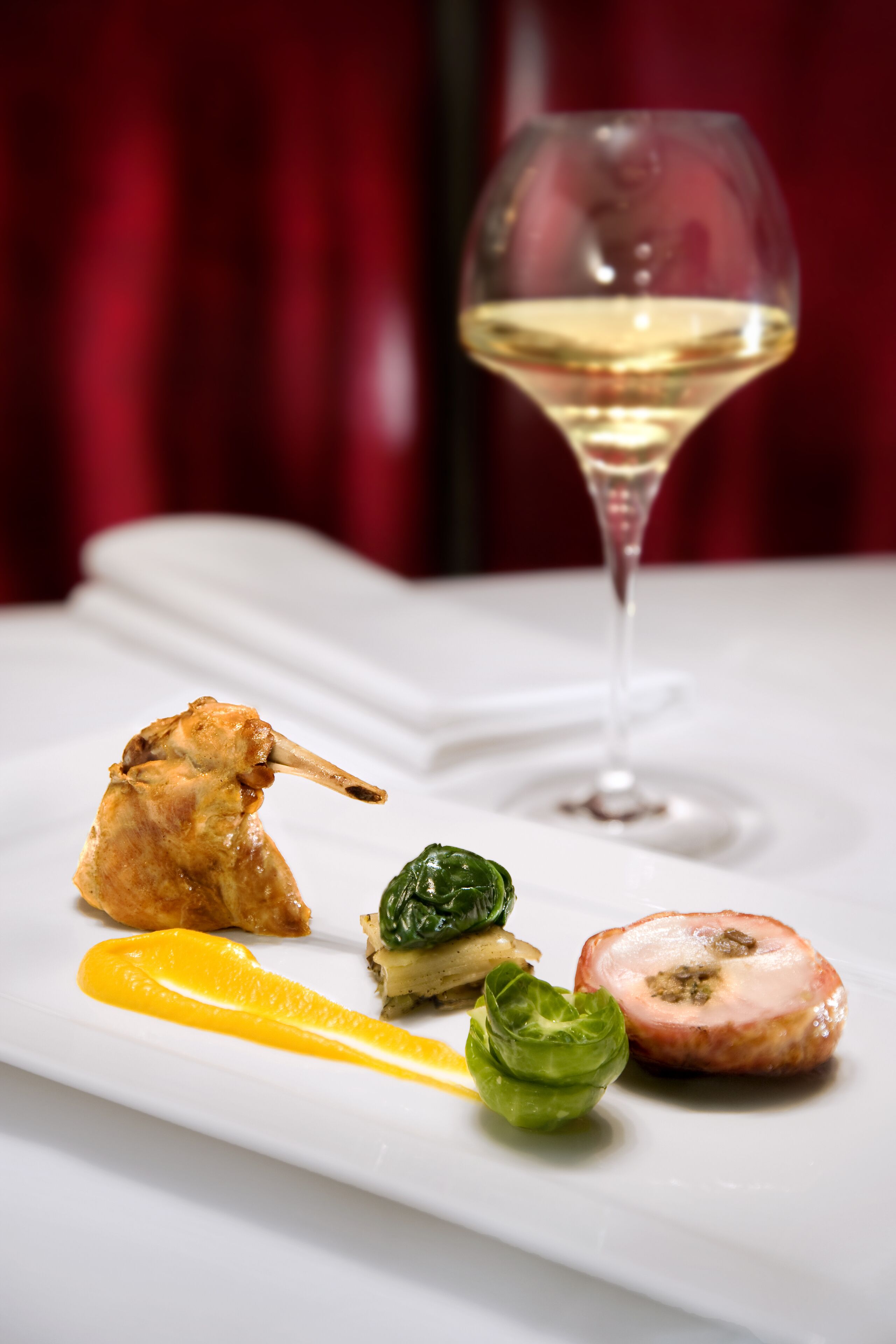 A white plate with a seared duck leg, a slice of tomato, a basil leaf, and a slice of meat with a glaze, accompanied by a glass of white wine.