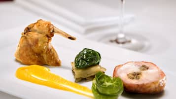 A white plate with a seared duck leg, a slice of tomato, a basil leaf, and a slice of meat with a glaze, accompanied by a glass of white wine.