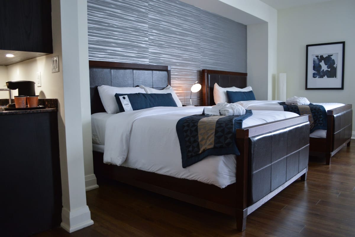 A hotel room with two beds, a wooden headboard, a large window with blinds, a framed artwork on the wall, and a small table with a lamp.