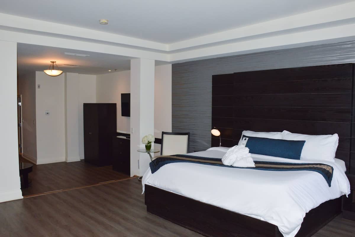 A modern hotel room with a large bed, a dark headboard, a bedside table with a lamp, and a view into another room.