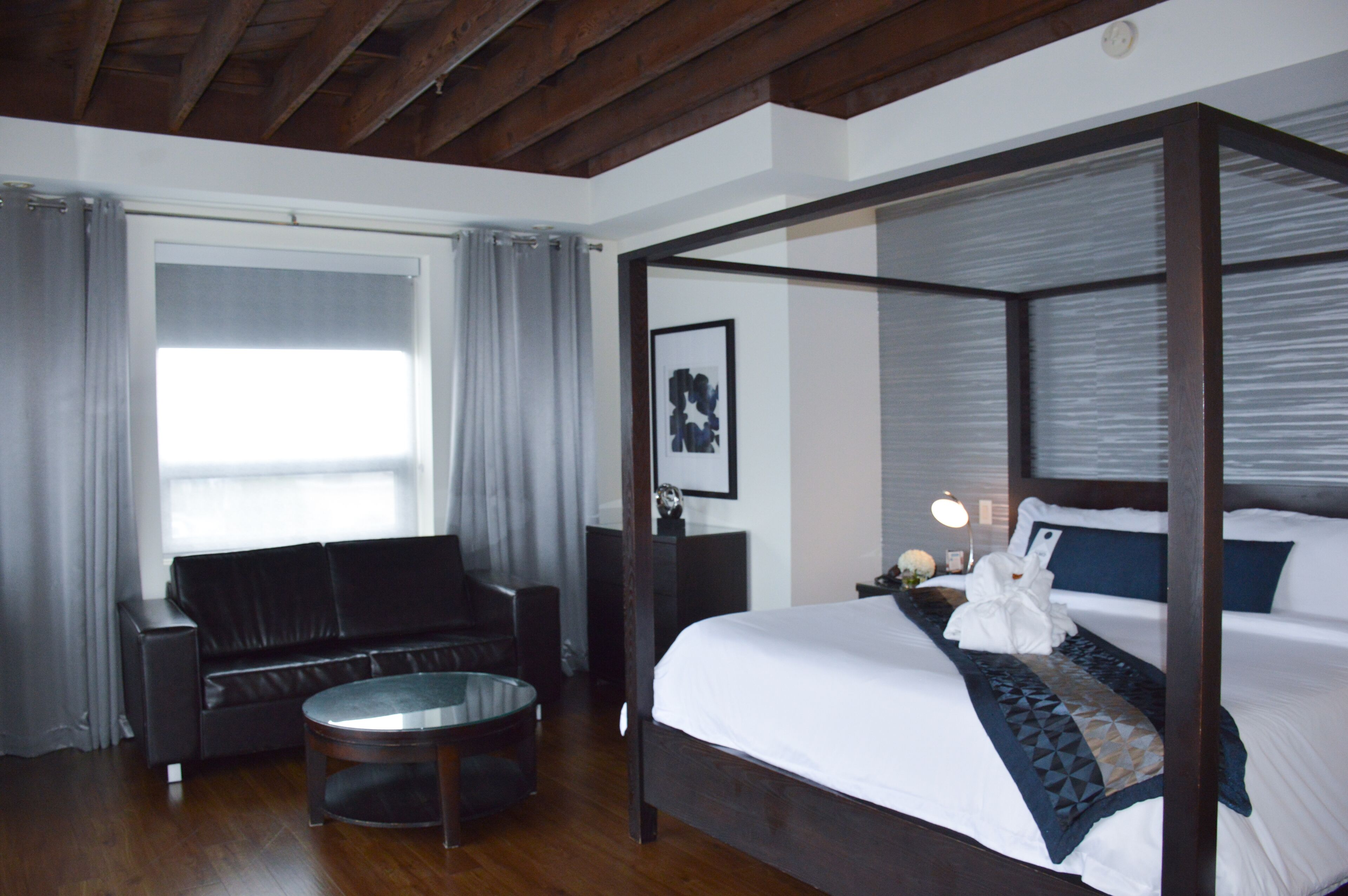 A four-poster bed with a white and blue bedspread, a round coffee table, a black leather sofa, and a large window with grey curtains.