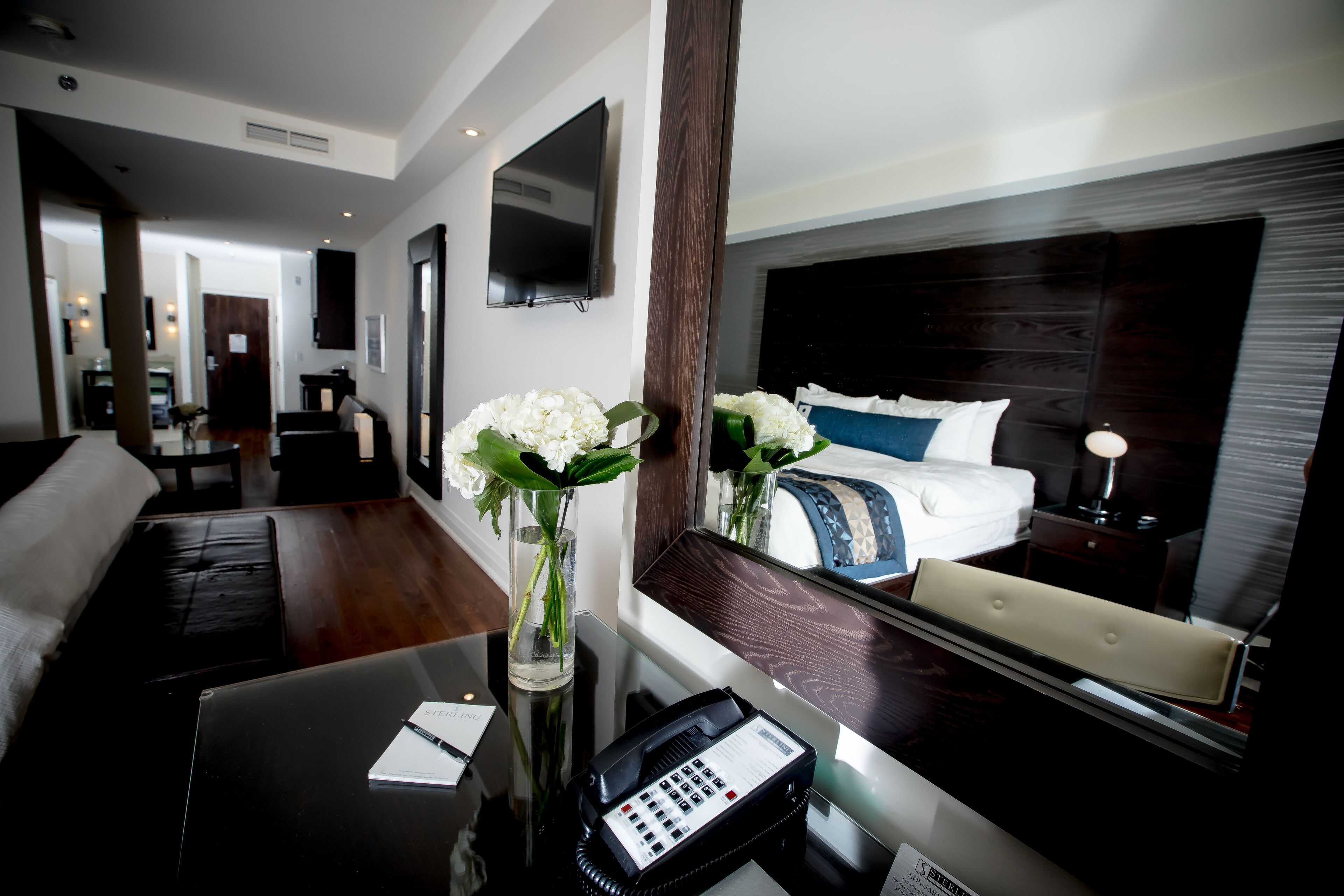 Habitación de hotel con cama, escritorio con teléfono, florero con flores y televisión montada en la pared.