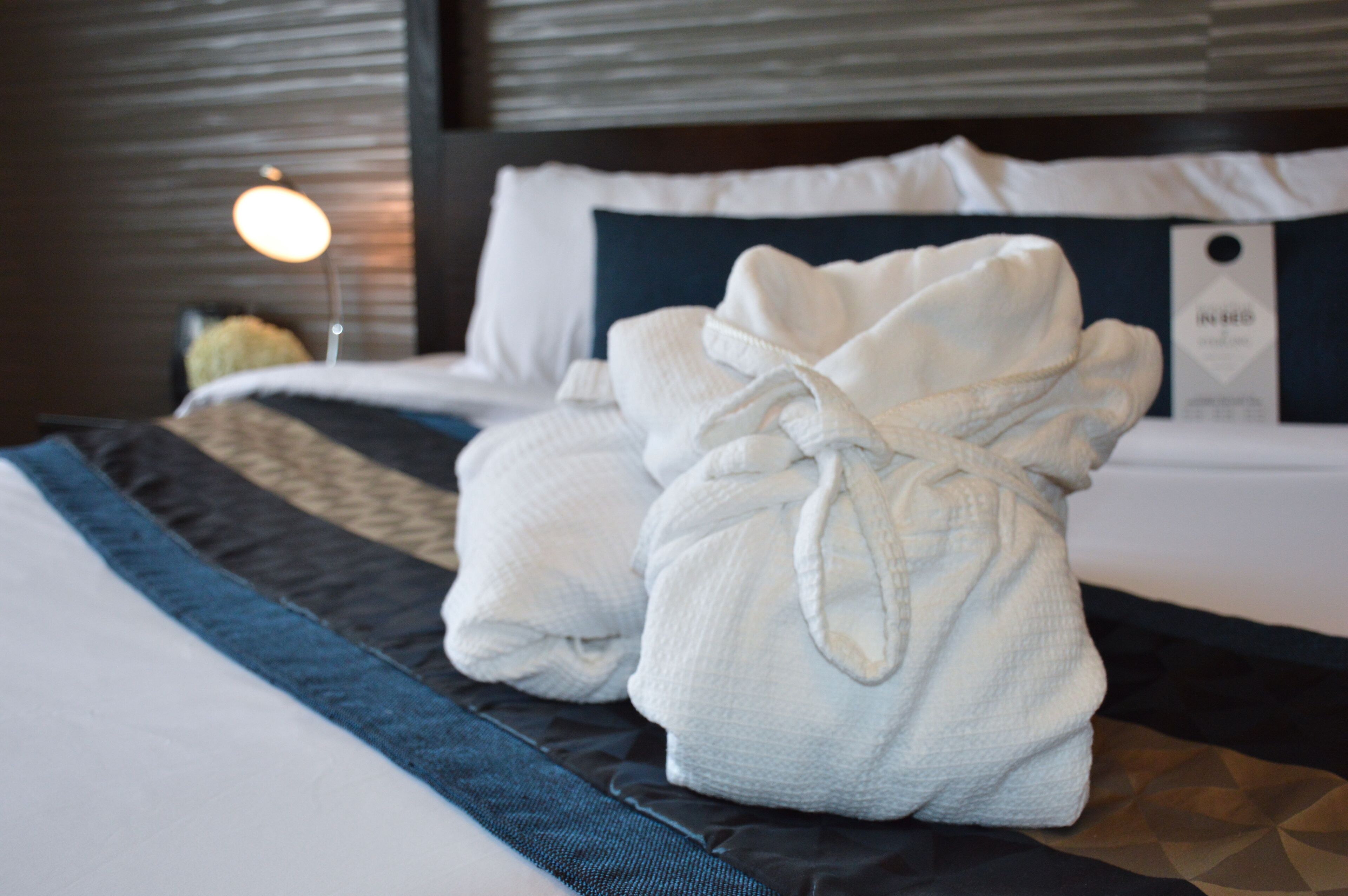 A neatly made bed with two white bathrobes tied at the ends, a dark blue and black striped pillow, and a white pillow with a dark blue accent.