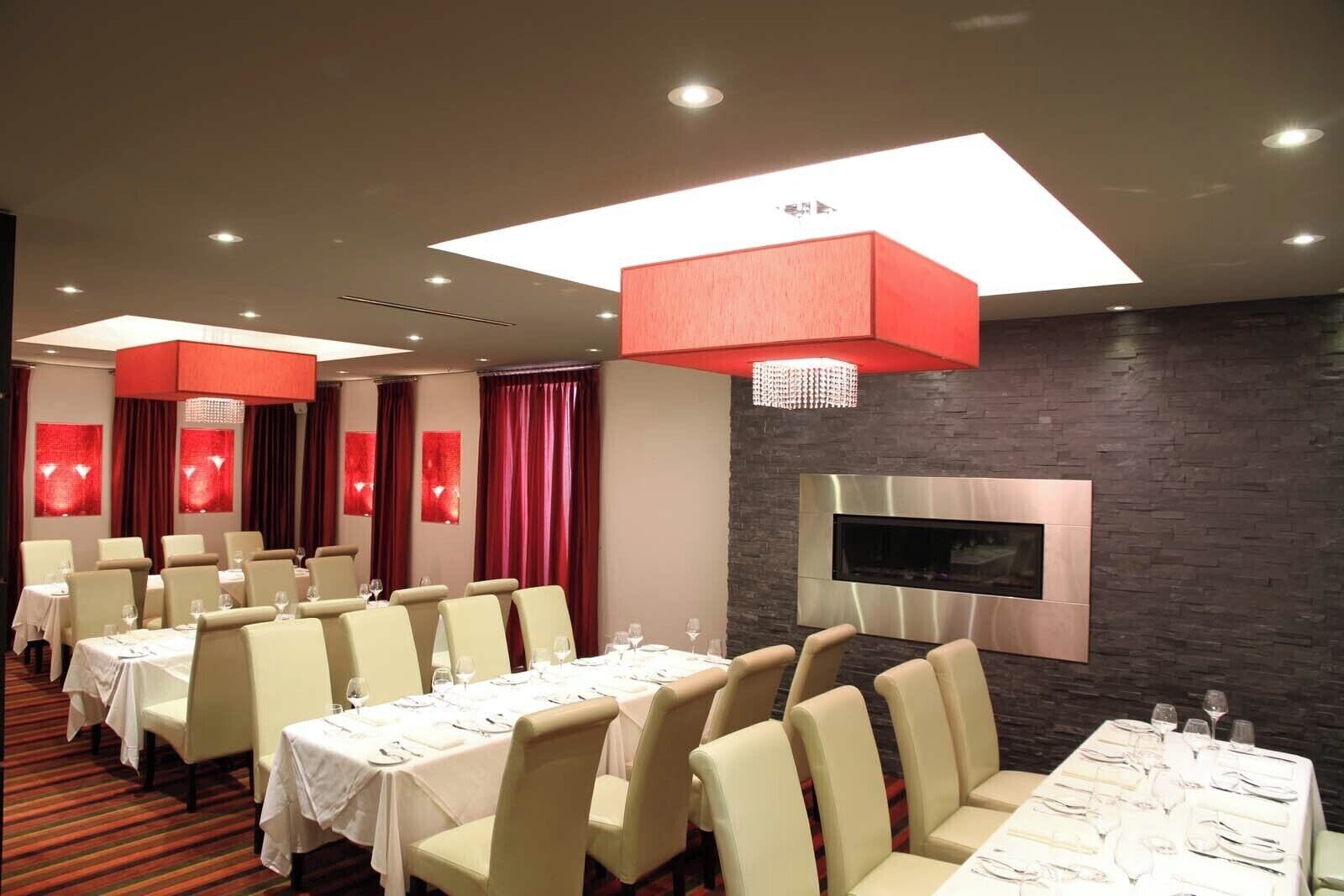 A modern dining area with multiple tables and chairs, a flat-screen TV mounted on a stone wall, and red curtains.