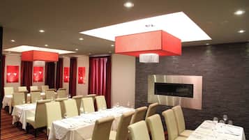 A modern dining area with multiple tables and chairs, a flat-screen TV mounted on a stone wall, and red curtains.